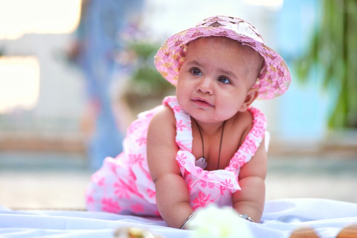 Cute baby girl in a pink floral dress and hat lying on a blanket outdoors.